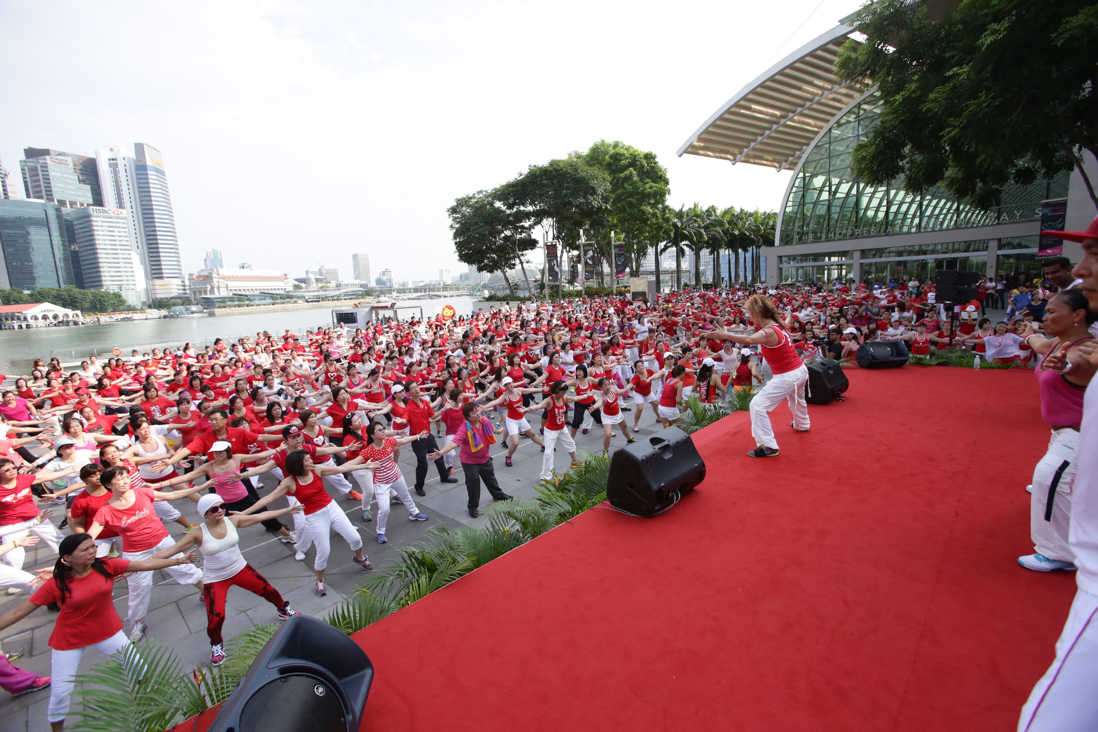 A large group of people in red and white outfits exercise outdoors near a waterfront in an urban area.